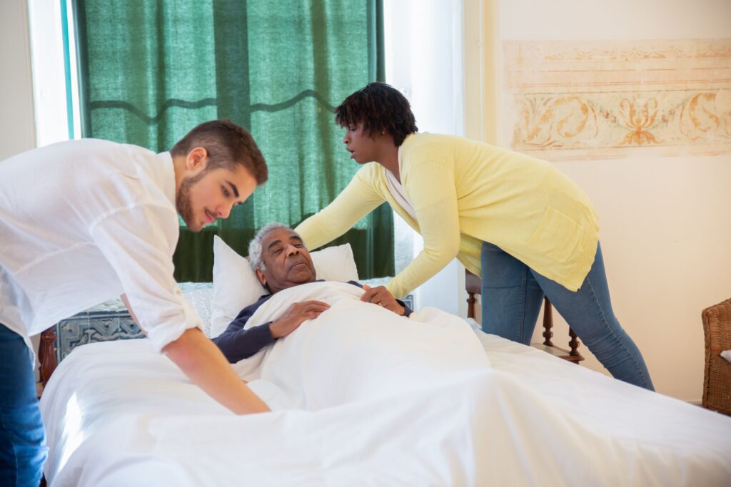 An elderly man resting in bed and assisted by two companions